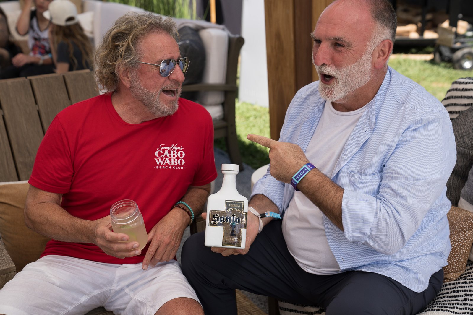 Two men laughing in a festival hospitality lounge, one holding a drink and the other a white tequila bottle, photographed by Tara Lenehan