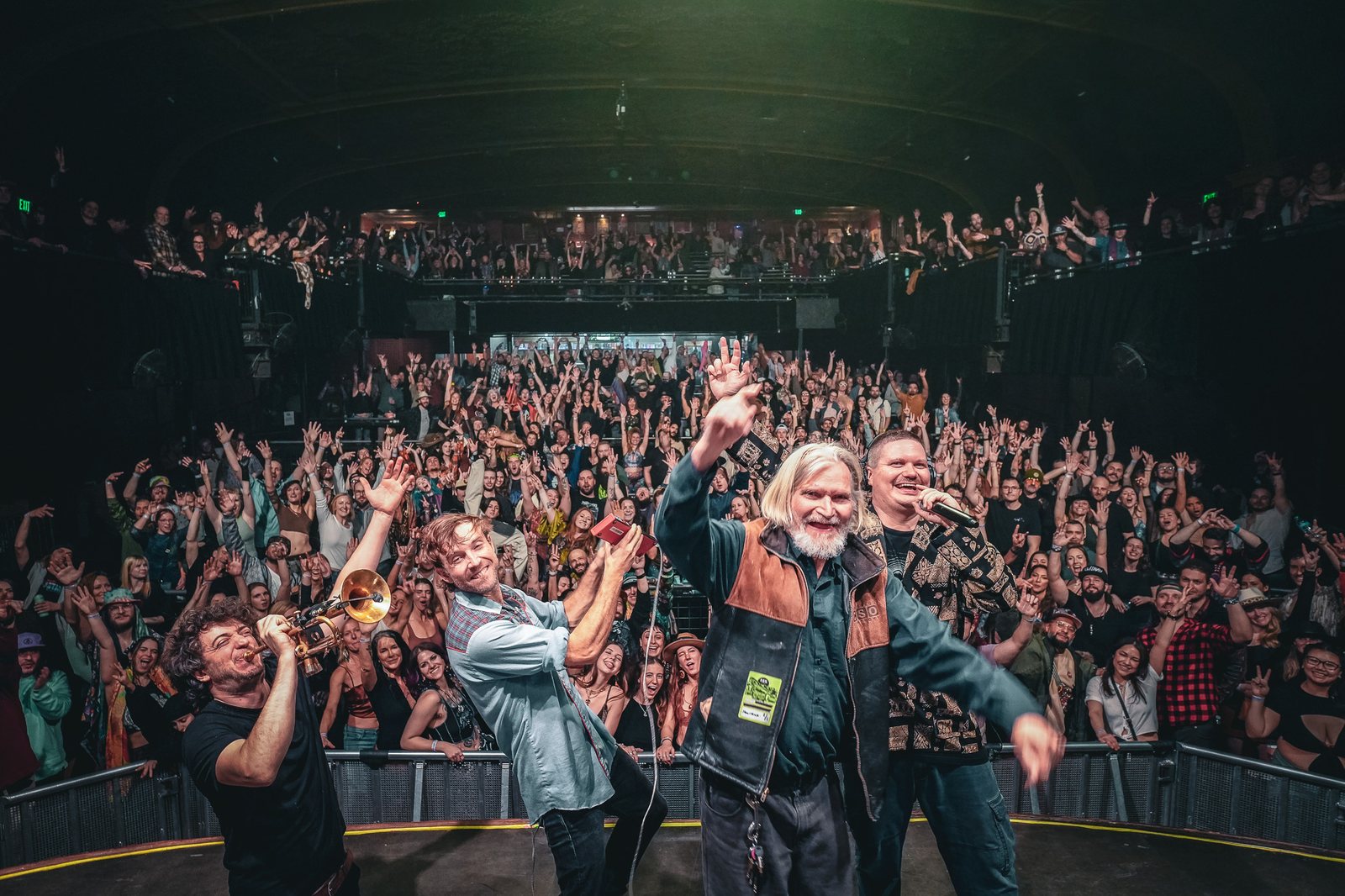 A band on stage waving and playing trumpet with a packed, cheering crowd behind them, photographed from the stage by live music photographer Tara Lenehan