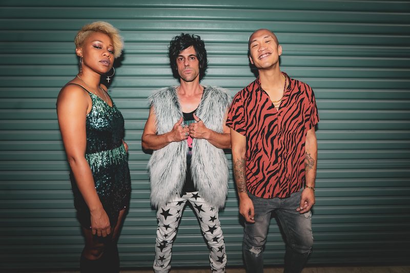 Press portrait of a three-piece band standing against a teal roll-up door