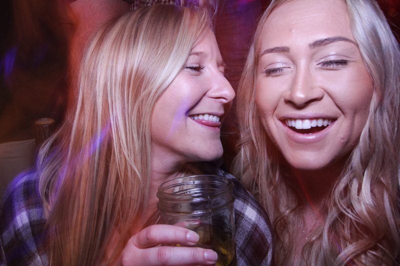 Two women laughing together over drinks in warm low light, lifestyle brand imagery