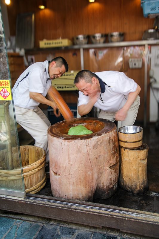 Two makers pounding rice with a wooden mallet behind a shop counter