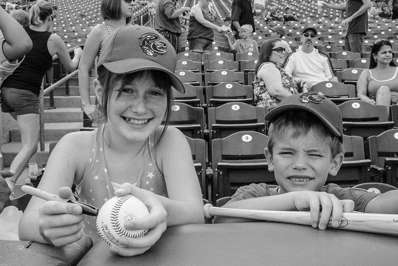 Two children leaning on a railing in the stands holding a baseball and bat