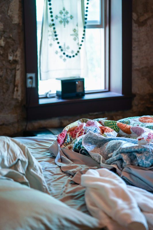 An unmade bed with layered quilts beneath a stone window in morning light