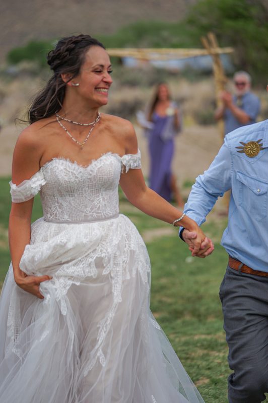 Bride in a lace wedding gown laughing while holding her groom's hand at a hot spring wedding in California