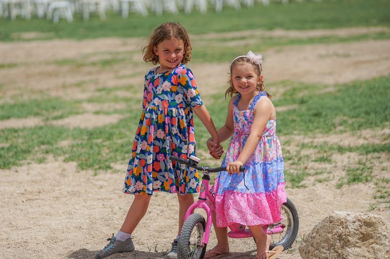 Two girls in colorful dresses with a pink bicycle in a grassy field at a wedding