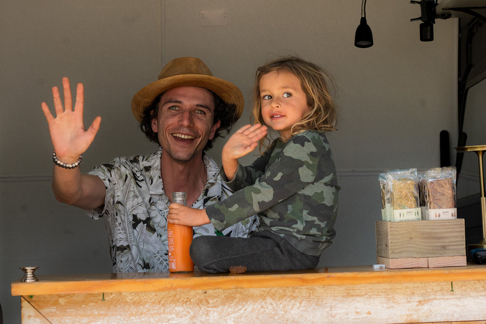 A dad in a straw hat and his young son waving from behind a wooden counter