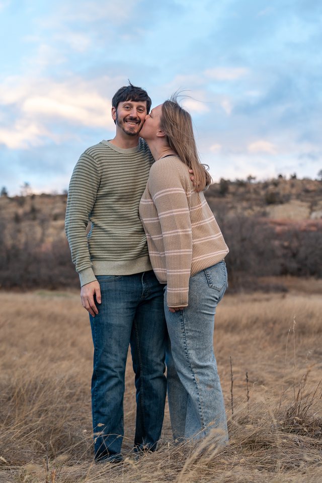 A woman kissing her grinning partner on the cheek in a windy autumn field
