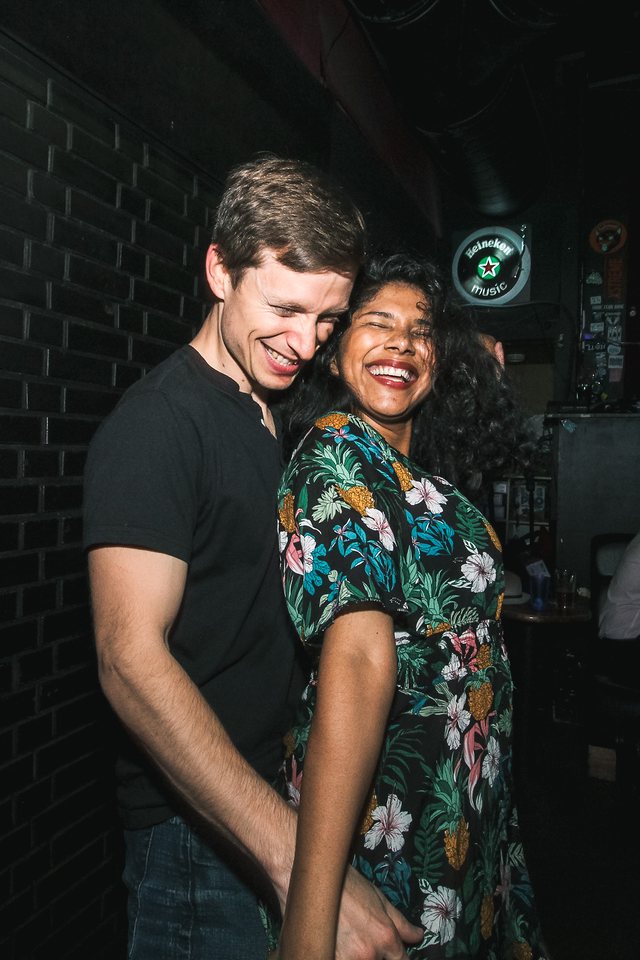 A couple laughing with their eyes closed, dancing close against a dark brick wall