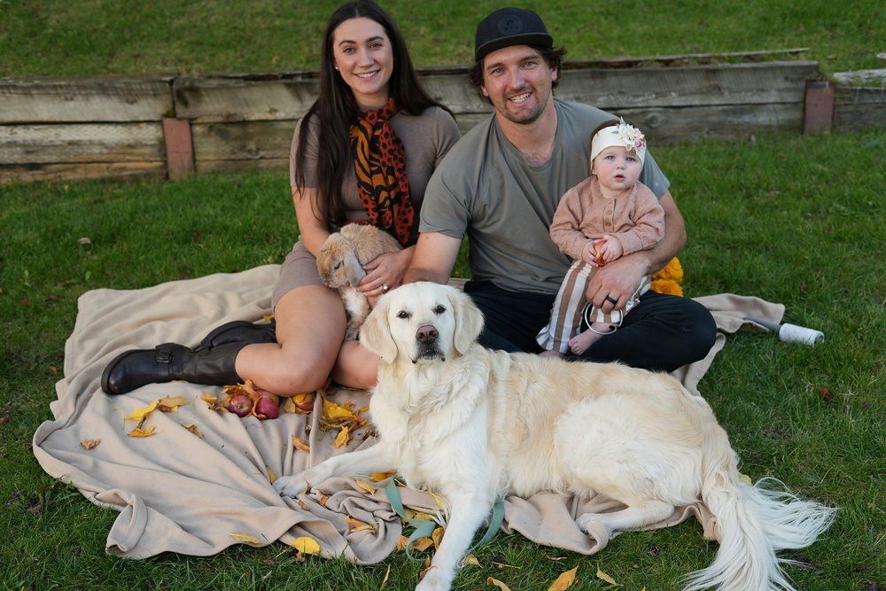 A family on a picnic blanket scattered with fall leaves and apples: mom holding a lop-eared bunny, dad holding their baby, and their golden retriever lying in front