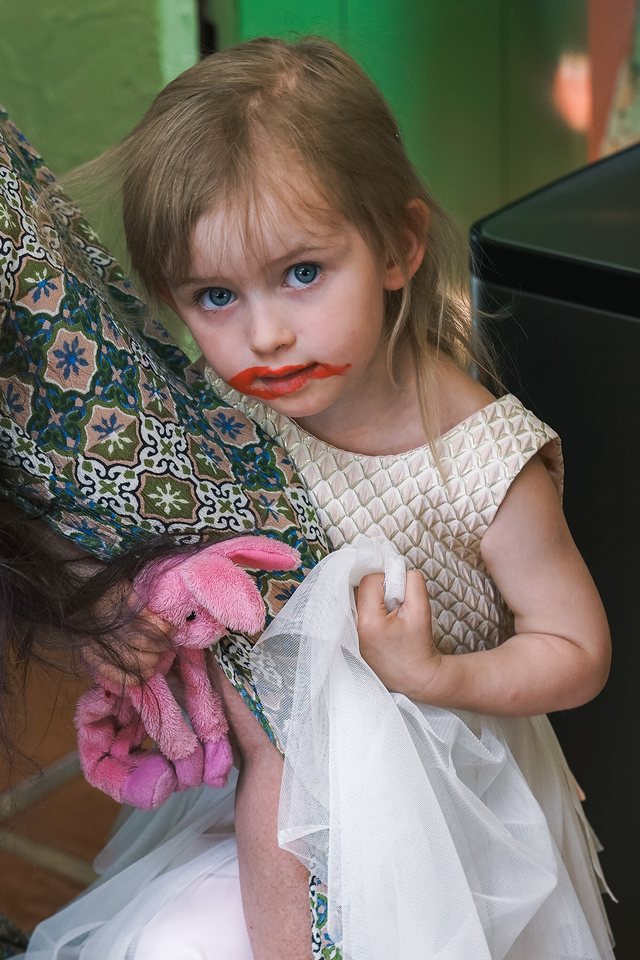 A little girl with lipstick smeared across her face peeking out from her mom's shoulder, holding a pink stuffed bunny