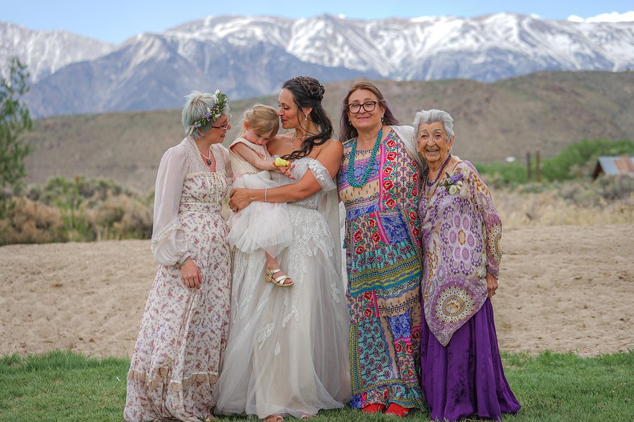 A bride holding a toddler, arm in arm with women of three generations in bright dresses, snowy mountains behind them