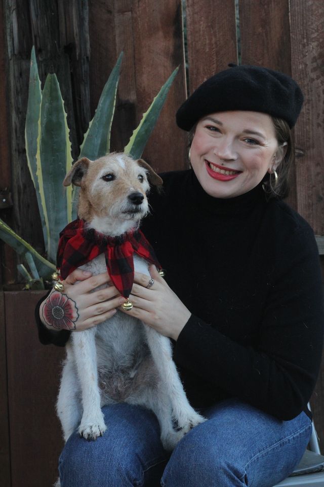A smiling woman in a black beret holding her grey-muzzled terrier, dressed in a plaid holiday scarf with jingle bells