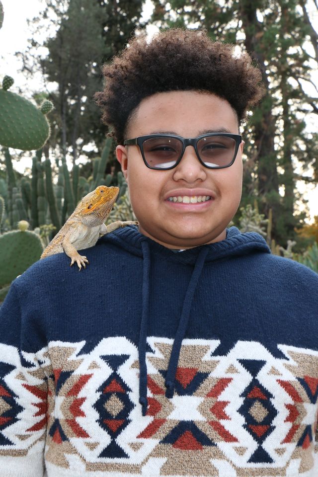 A smiling teen in glasses with his bearded dragon perched on his shoulder in a cactus garden