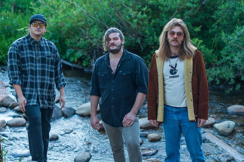 Three members of Rainy Eyes stand in a rocky creek bed surrounded by evergreens near Winter Park