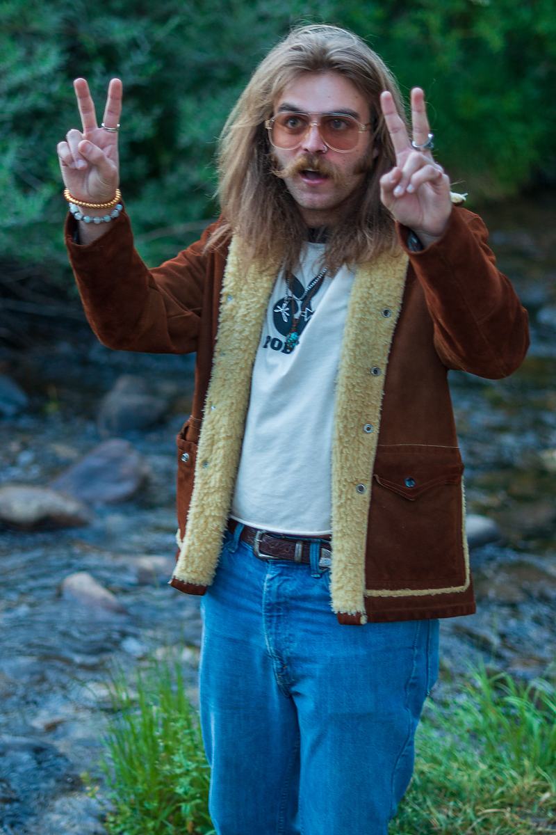 A Rainy Eyes musician in a corduroy jacket throws double peace signs while standing in a creek, a band portrait