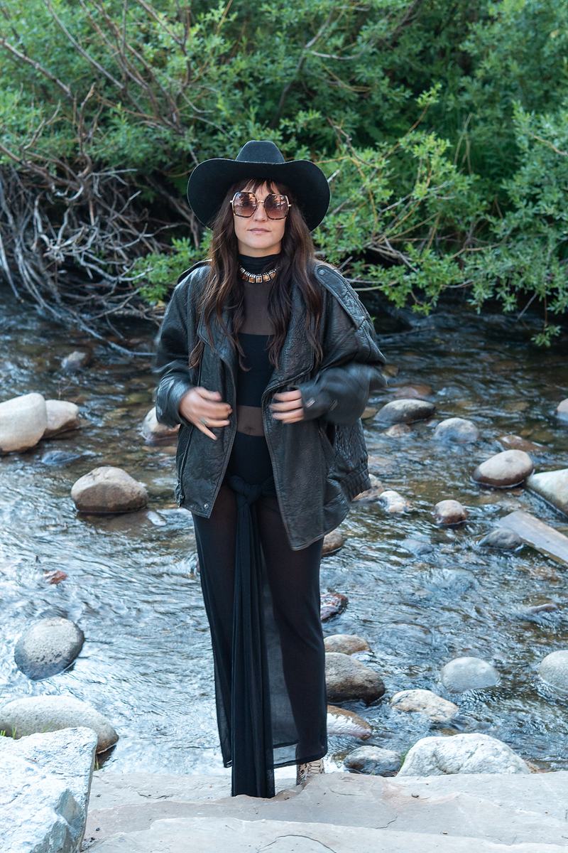 The vocalist of Rainy Eyes stands beside a mountain creek in a black hat and long coat, a Winter Park portrait