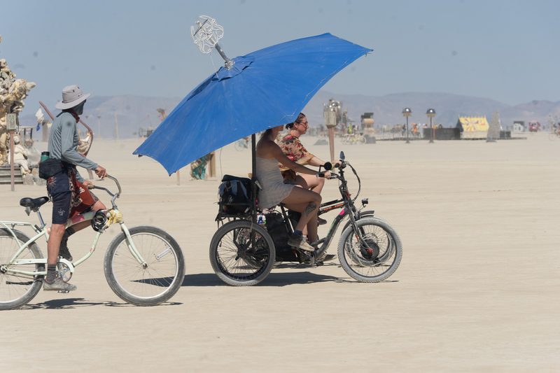 Cyclist with a blue umbrella and another rider on motorized bikes across the open playa at Burning Man