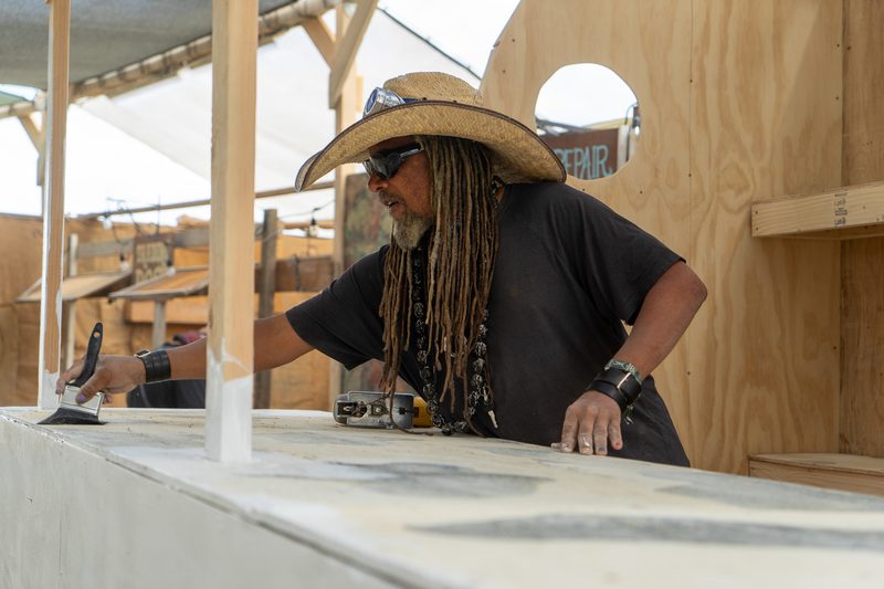 Builder in a straw hat and dreadlocks working on a plywood frame at Burning Man