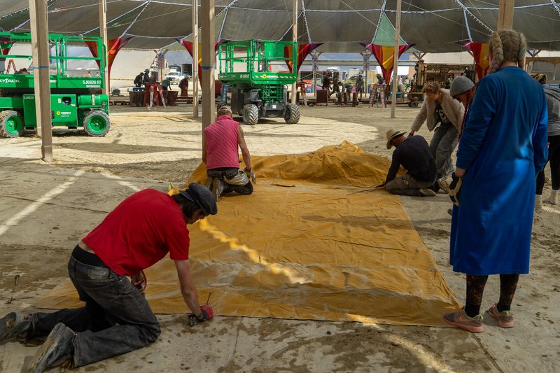 Crew working on a yellow tarp beneath a large shade structure with lifts in the background at Burning Man