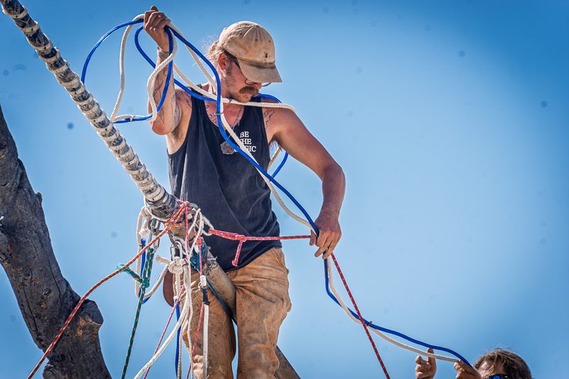 Climber rigging ropes and cables high on a pole against a bright blue sky at Burning Man