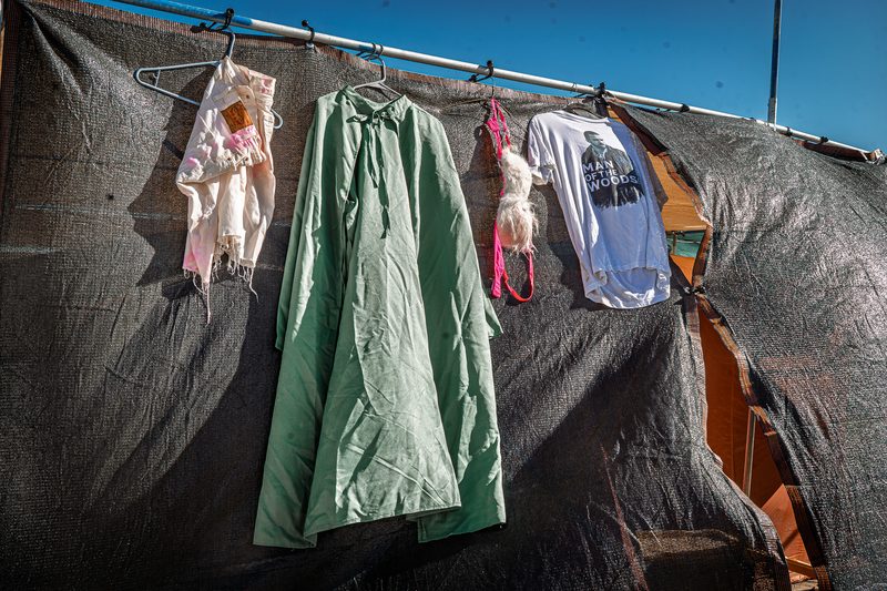 Colorful clothes and a green cape hanging on a dark camp wall under a blue sky at Burning Man