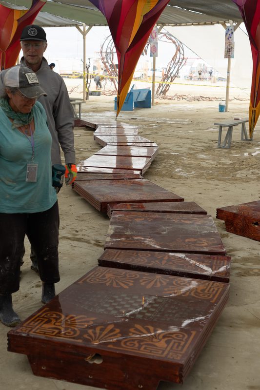 Crew members arranging large rust-colored wooden panels on the playa under a shade structure at Burning Man