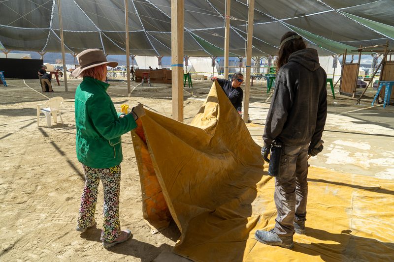 Two crew members in colorful clothes handling a large yellow tarp inside a shade structure at Burning Man