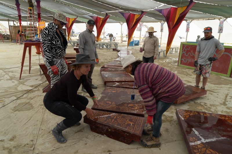 Crew members lifting large red wooden panels under a shade structure at Burning Man