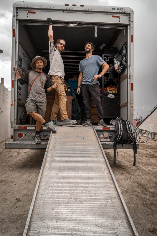 Three crew members posing at the open back of a box truck at a ramp at Burning Man