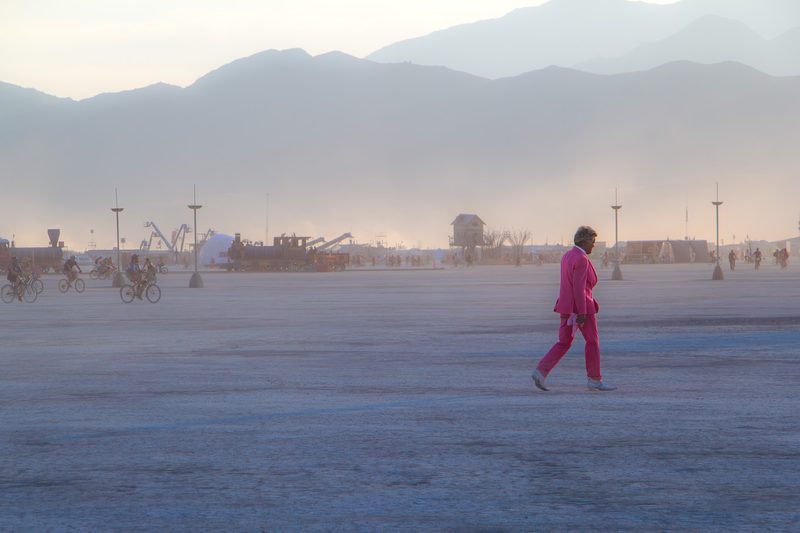 Lone figure in pink walking across the hazy playa toward distant mountains and art at Burning Man