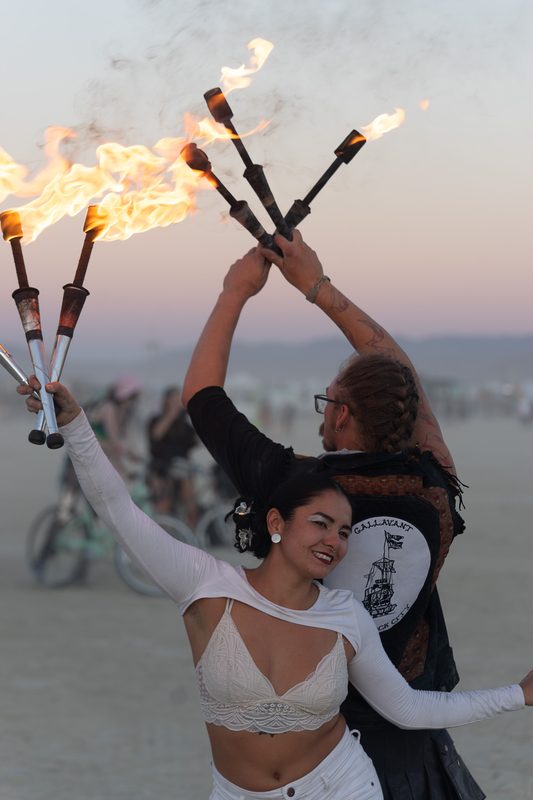Two fire performers crossing flaming staffs at dusk on the playa at Burning Man