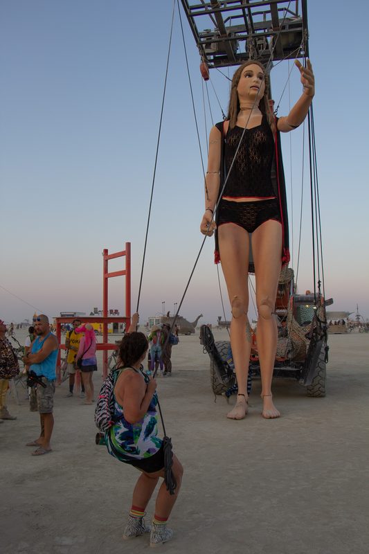 Giant standing figure hanging from a crane with visitors below at dusk at Burning Man