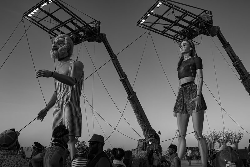 Black and white view of giant human figures raised on cranes above a crowd at Burning Man
