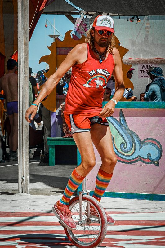 Man in a bright red outfit riding a unicycle past a colorful camp at Burning Man