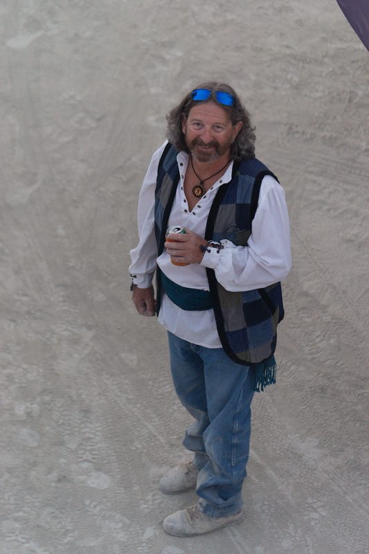 Man in a vest and blue jeans standing on pale cracked playa ground at Burning Man
