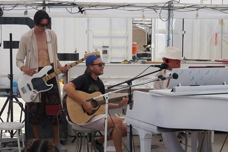 Musicians playing guitar and bass beside a white piano in a camp at Burning Man