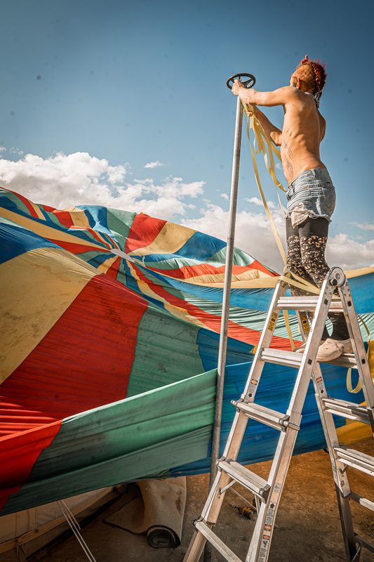 Person standing on a ladder adjusting a colorful striped canopy against the sky at Burning Man