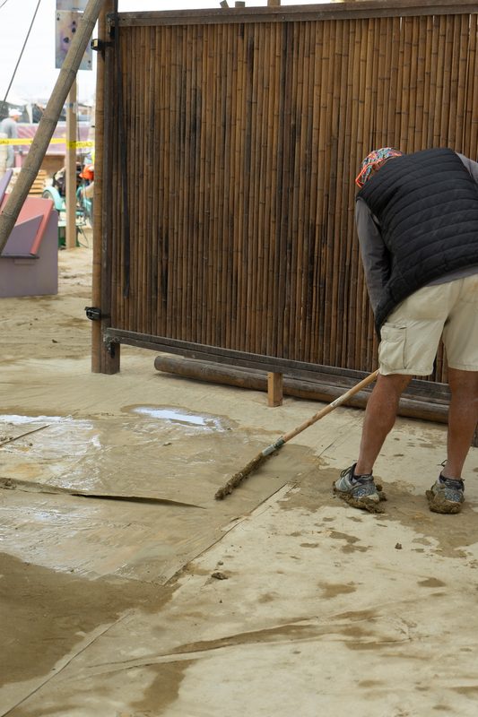 Person sweeping playa dust from a floor beside a tall bamboo wall at Burning Man