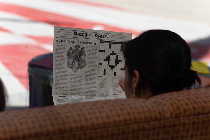 Person reading a newspaper crossword from a couch with a red and white pattern behind at Burning Man