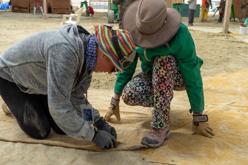 Two crew members kneeling to secure material on the dusty playa floor at Burning Man