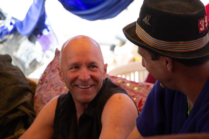 Bald man smiling at the camera beside a friend in a brimmed hat inside a colorful camp at Burning Man