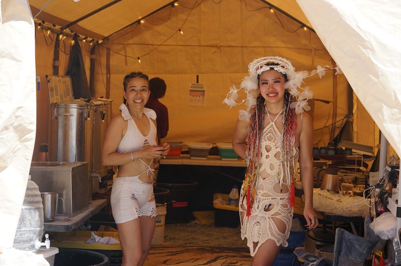 Two women in white outfits standing in a warmly lit camp tent at Burning Man