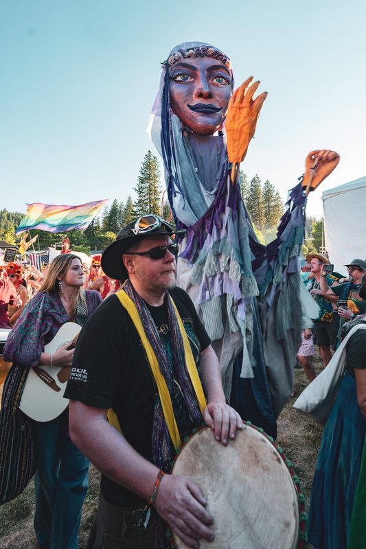 Giant puppet in a hooded costume beside a hand drummer in a festival crowd