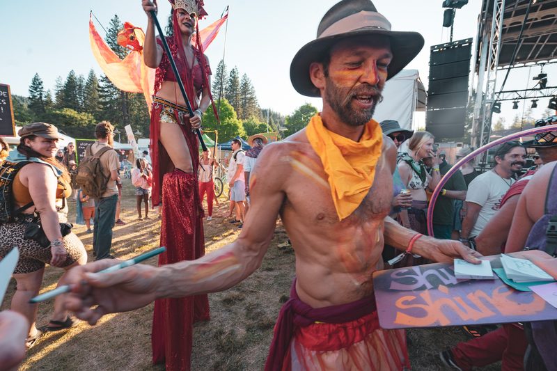 Performer in a yellow bandana beside a stilt walker in costume at a festival