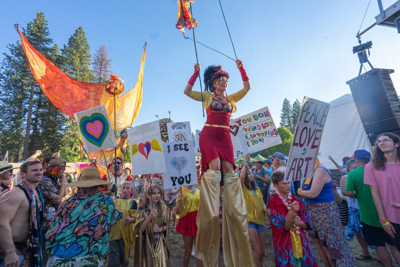 Stilt walker in a colorful costume leading a festival parade past signs and crowds