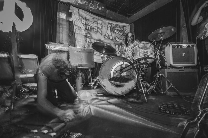 Black and white photo of a performer crouched at the front of the stage near a drum kit during an Insects vs. Robots show