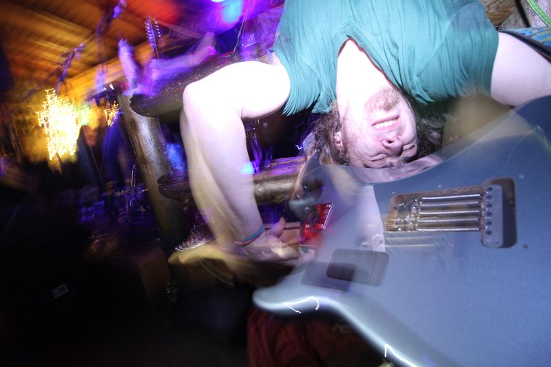 Color photo of a guitarist with teal hair bent over his guitar in purple stage light during an Insects vs. Robots show