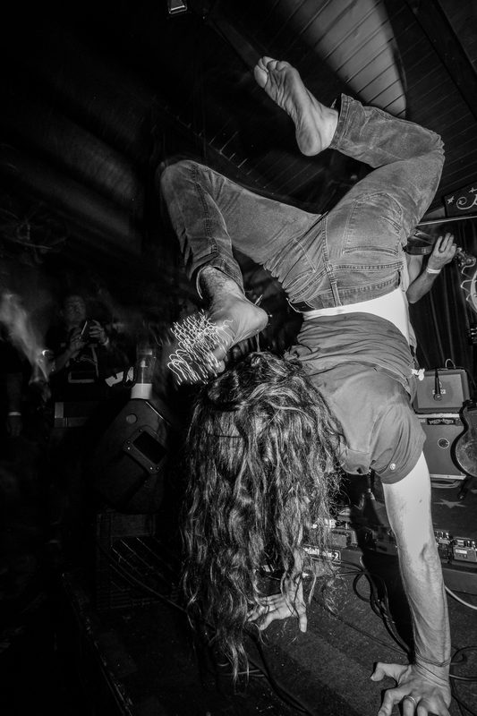 Black and white photo of a performer upside down on stage with long hair hanging during an Insects vs. Robots show